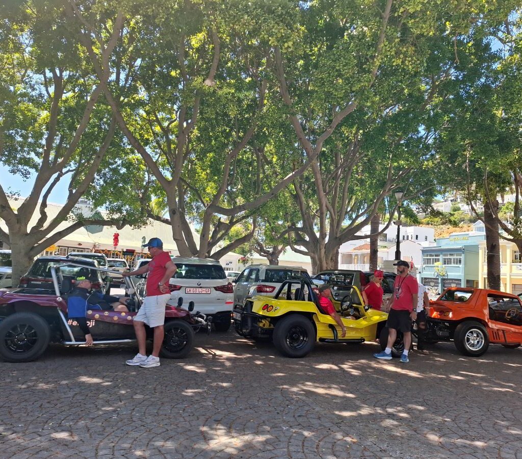 Wild Buggers Beach Buggy Club members exploring the stalls and the Just Nuisance statue on the shores of Simon's Town during their Valentine's Day run. Classic VWs parked nearby.