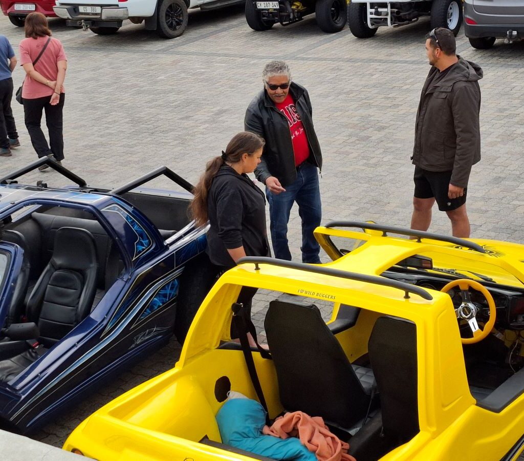 Wild Buggers Beach Buggy Club members, all wearing something black as a tribute, standing casually and chatting in the Potbelly Pantry parking area, with various beach buggies and air-cooled VWs visible in the background during Wiehann's Run.