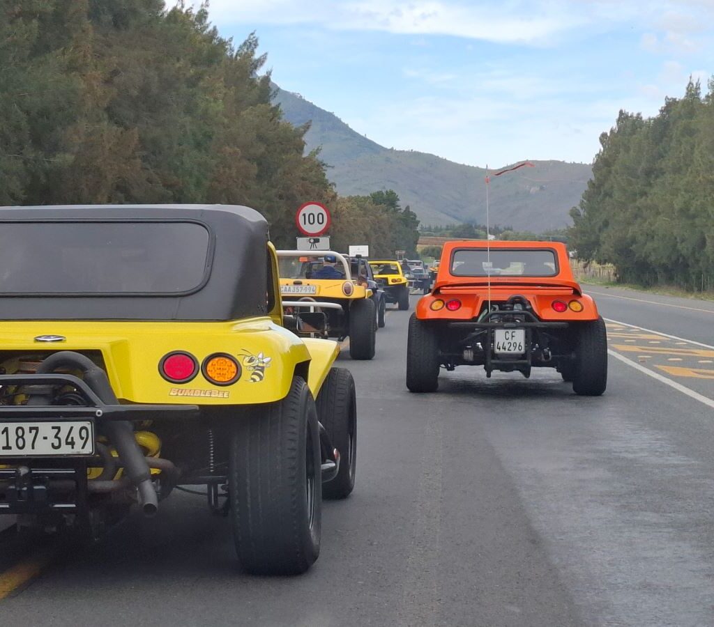 A convoy of diverse Wild Buggers beach buggies and air-cooled VWs, driving along a scenic road under a clear sky, heading towards their next destination on Wiehann's Run.