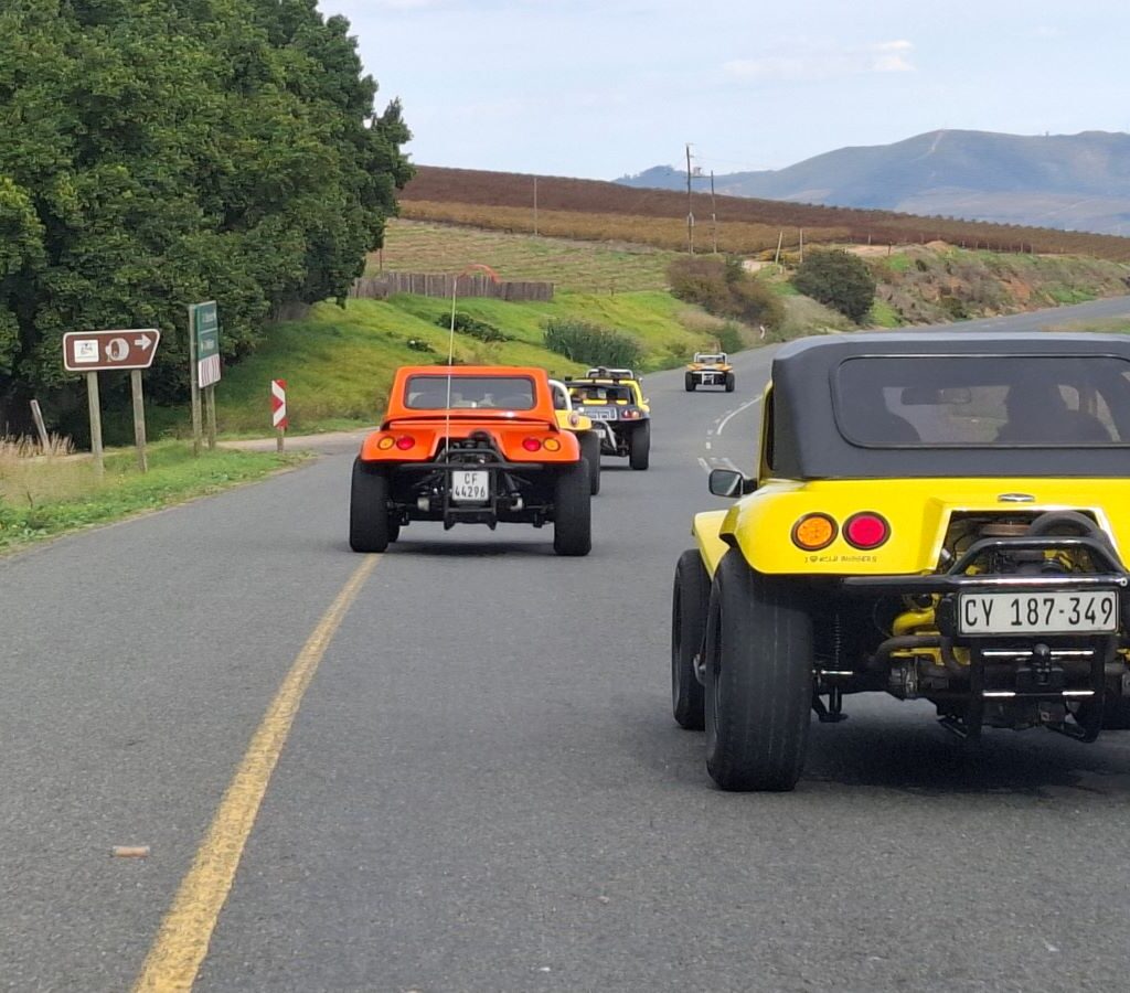 A group of Wild Buggers beach buggies and various air-cooled VW vehicles driving in a relaxed convoy along a winding, scenic road bordered by lush greenery and distant hills, showcasing the beautiful South African landscape during Wiehann's Run.