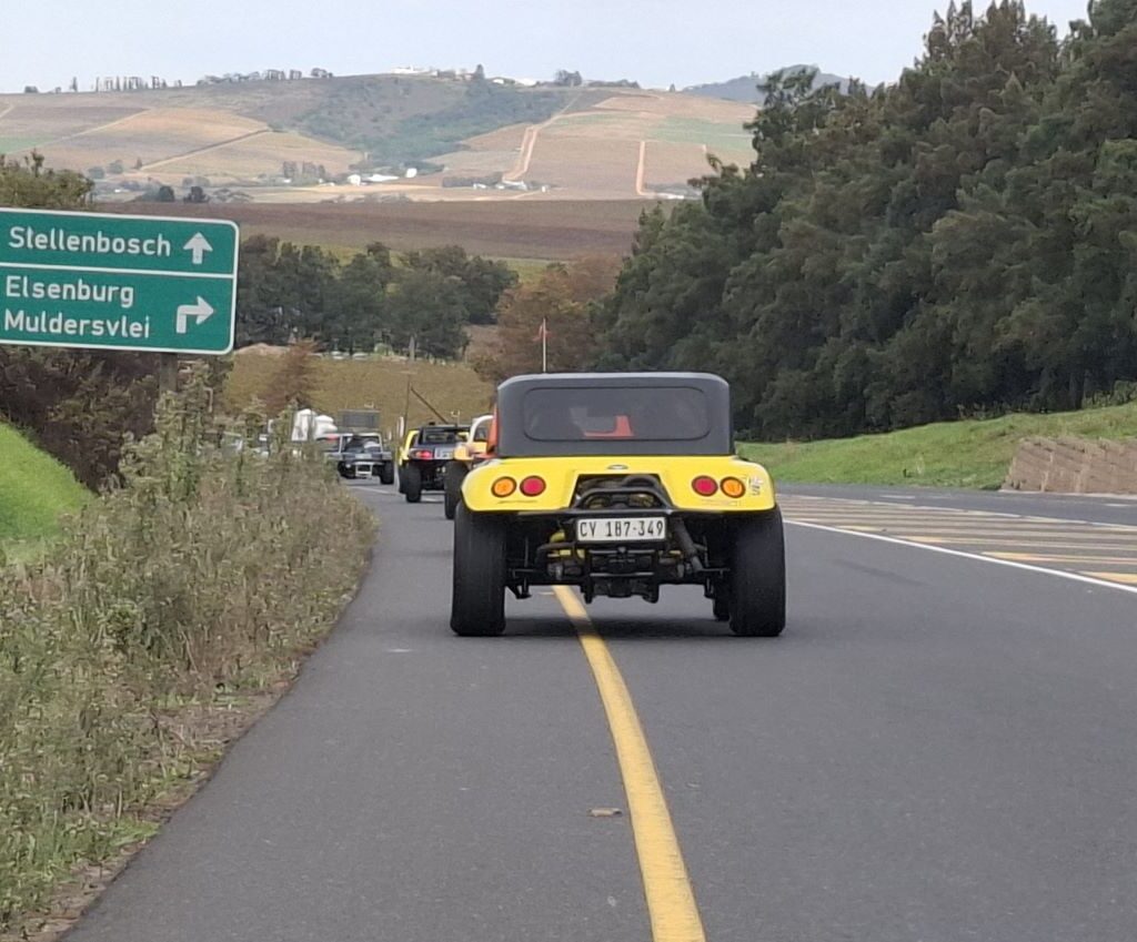 A convoy of Wild Buggers beach buggies and various air-cooled VW vehicles driving past a road sign indicating the turn-off for Elsenburg and Muldersvlei, under a clear South African sky, during Wiehann's Run.