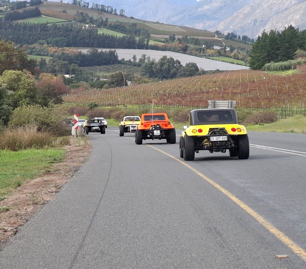 A long line of Wild Buggers beach buggies and various air-cooled VW vehicles driving in a tight convoy along a winding road, surrounded by expansive, vibrant green hills and clear blue skies, highlighting the beautiful South African scenery during Wiehann's Run.