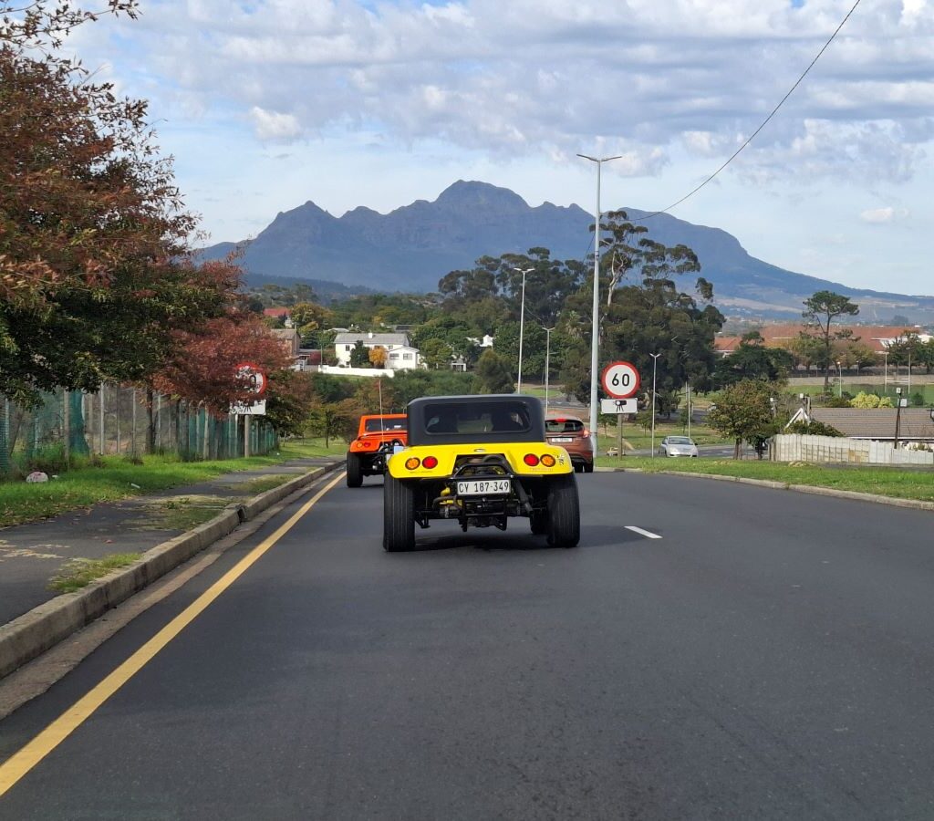 A convoy of Wild Buggers beach buggies and various air-cooled VW vehicles driving into a small, welcoming town, with local buildings and street signs visible, indicating the group's arrival at a new destination during Wiehann's Run.