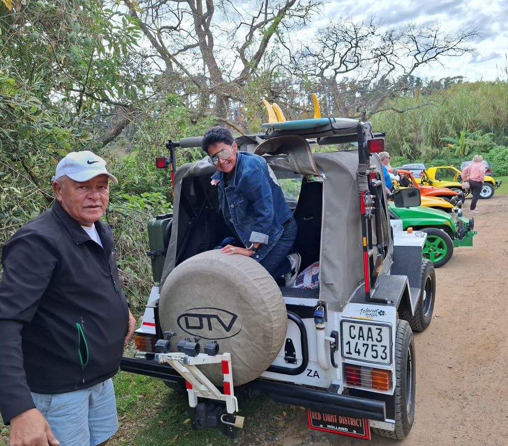 Faizal gently helps Ayesha as she exits their "Veep" beach buggy in the parking area of Helderberg Farm during Wiehann's Run, showing a moment of care and camaraderie.