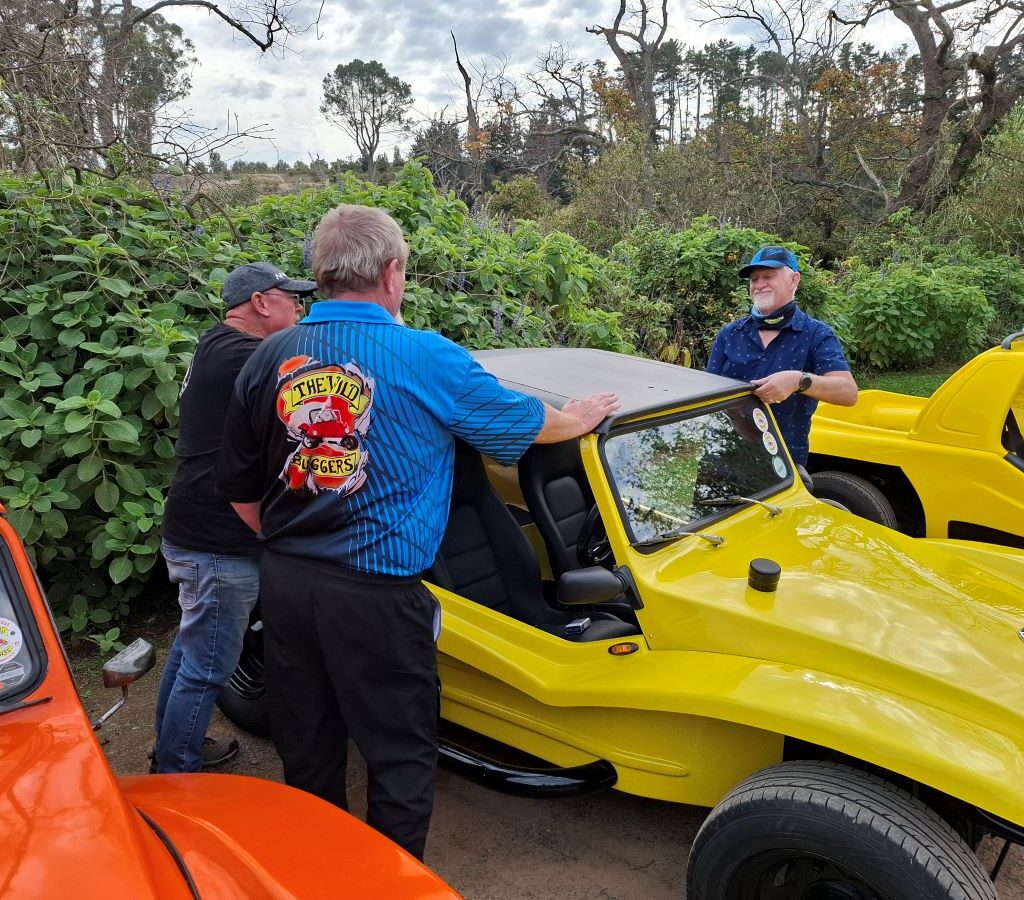 Chris, Anthony, and Jacques from the Wild Buggers Beach Buggy Club stand together in the Helderberg Farm parking area, engrossed in conversation and gesturing towards their beach buggies, all wearing something black as a tribute during Wiehann's Run.