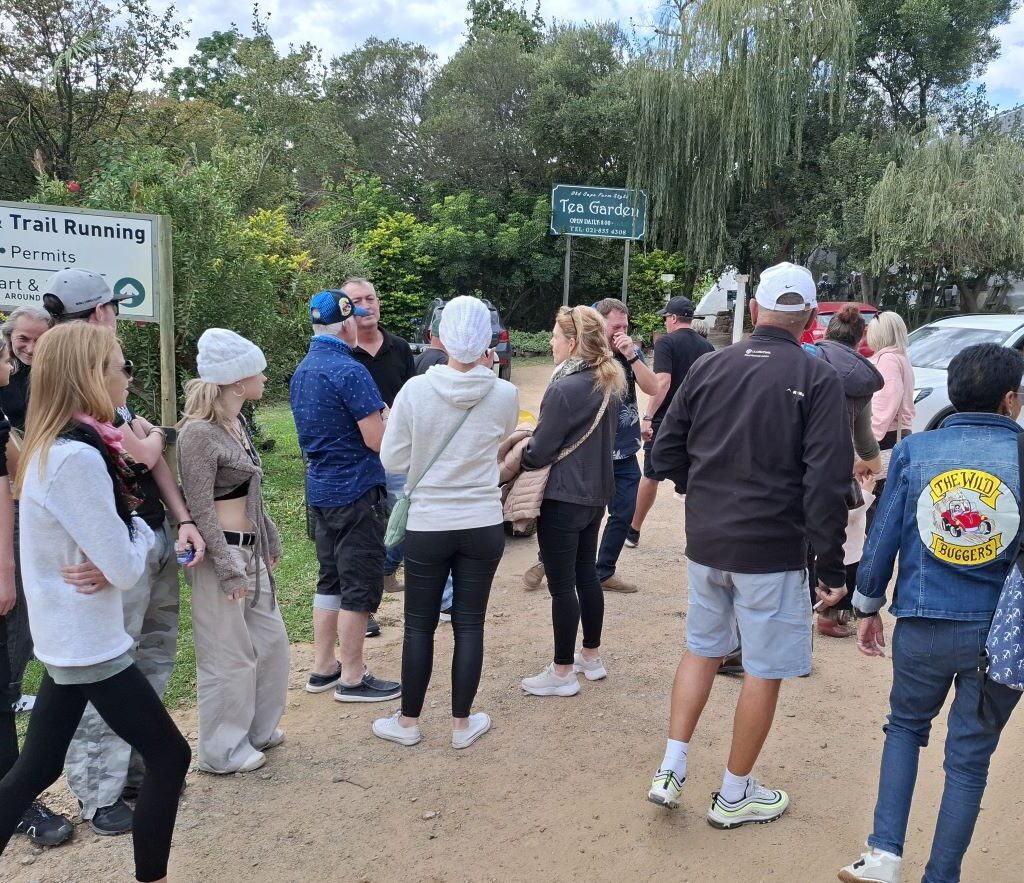 Anton Kleyn, President of the Wild Buggers Beach Buggy Club, stands among parked beach buggies in the Helderberg Farm parking area, gesturing and encouraging club members, who are chatting leisurely, to move towards the milkshake stop during Wiehann's Run.