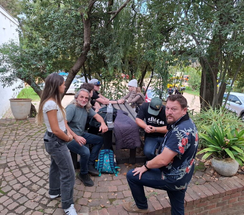 A group of newer Wild Buggers Beach Buggy Club members, wearing black as a tribute, seated at a table in the garden area of Helderberg Farm, engaged in conversation while patiently waiting for their milkshakes during Wiehann's Run.