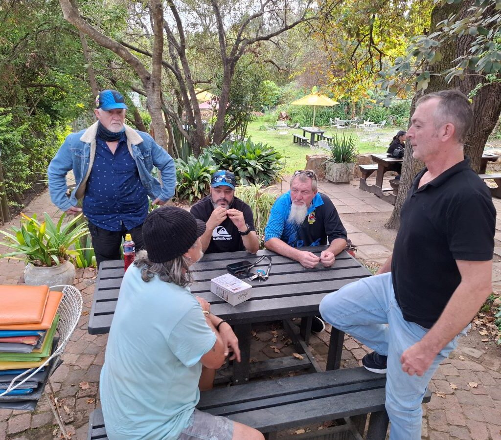 Wild Buggers Beach Buggy Club members are patiently waiting in the beautiful garden setting of Helderberg Farm, with lush greenery and scenic elements visible, while anticipating their milkshakes during Wiehann's Run.