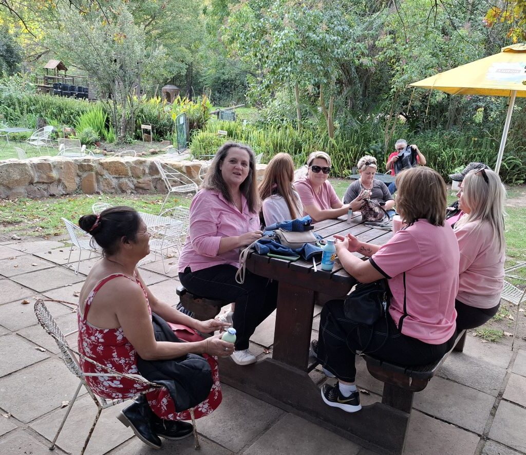 A group of Wild Buggers Beach Buggy Club ladies, all wearing black as a tribute, are patiently seated or standing in the beautiful garden area of Helderberg Farm, some smiling, as they continue to wait for their milkshakes during Wiehann's Run, suggesting a large group size.