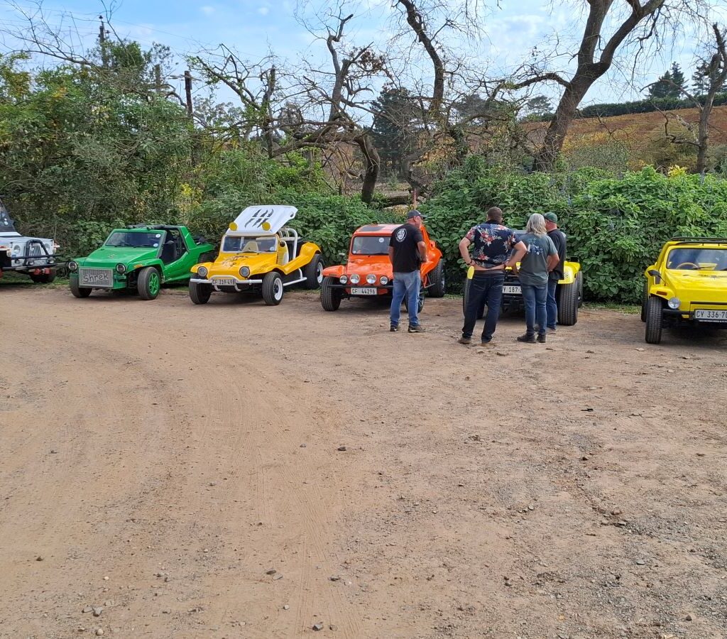 A group of Wild Buggers Beach Buggy Club members, wearing black as a tribute, are gathered in the Helderberg Farm parking area, engrossed in conversation around their parked beach buggies, seemingly drawn back to their vehicles despite being at the milkshake stop during Wiehann's Run.