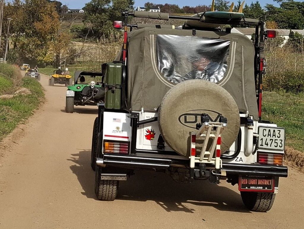 A convoy of brightly colored beach buggies kicking up dust as they embark on a scenic gravel road adventure near Helderberg Farm, with the majestic Helderberg Mountains in the background.