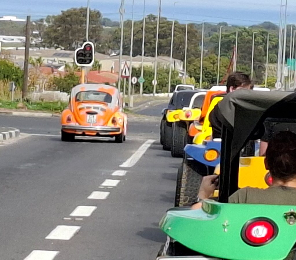 A line of Wild Buggers beach buggies stopped at a "robot" (traffic light) in South Africa, with a bright orange VW Beetle driving past, during Wiehann's Run.