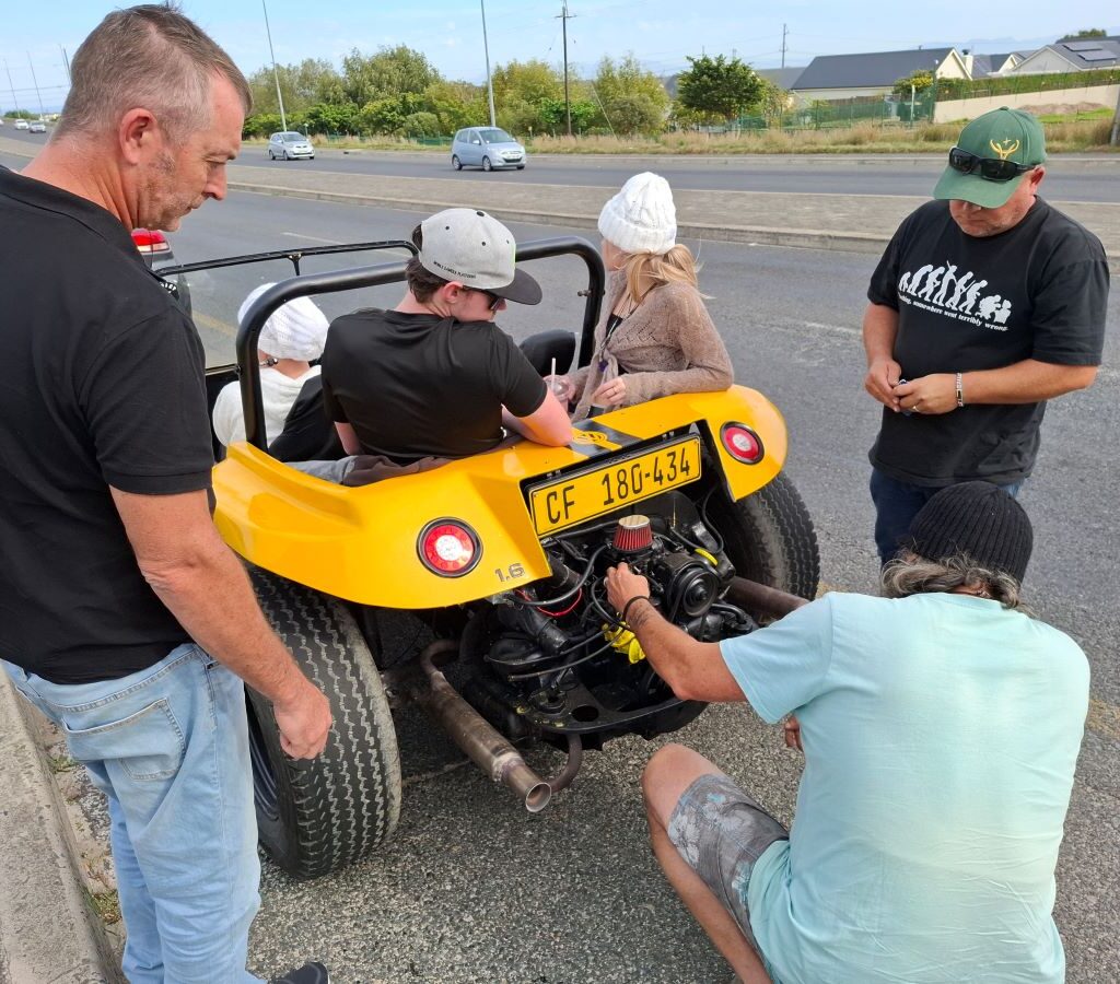 Tony and Rhoodie are kneeling and inspecting the engine of a stationary beach buggy on the side of a road in a scenic area of the Western Cape during Wiehann's Run, with other beach buggies from the Wild Buggers convoy parked nearby.