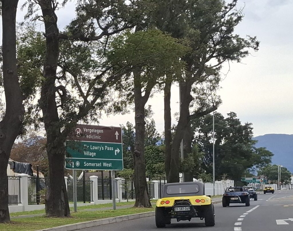 A convoy of Wild Buggers beach buggies is seen driving past the sign for Sir Lowry's Pass Village, on a scenic road, with the village and surrounding landscape visible in the background.