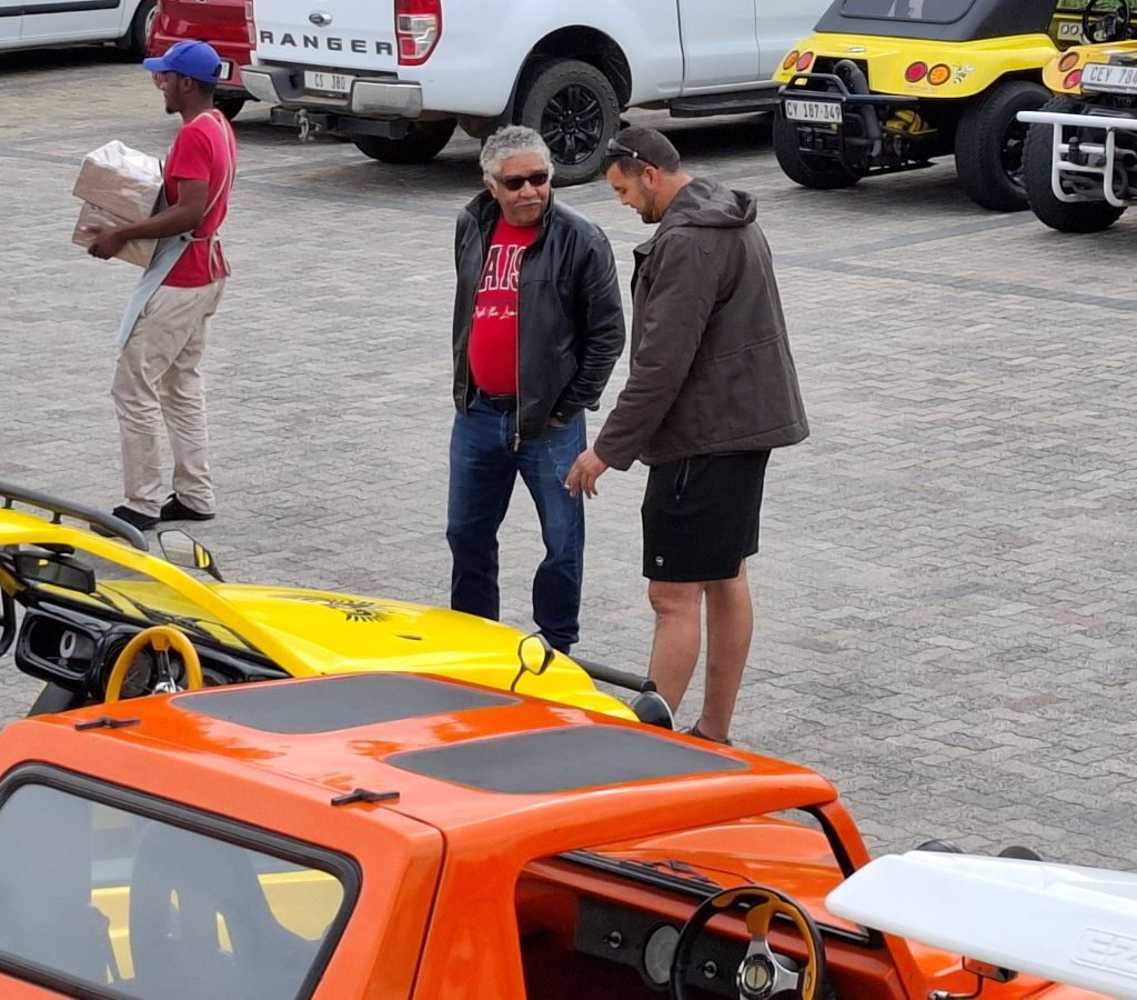 Members of the Wild Buggers Beach Buggy Club, gathered in the parking area at Potbelly Pantry in Klapmuts, with several parked beach buggies, air-cooled VWs, and classic cars visible in the background, chatting and enjoying the first stop of Wiehann's Run.