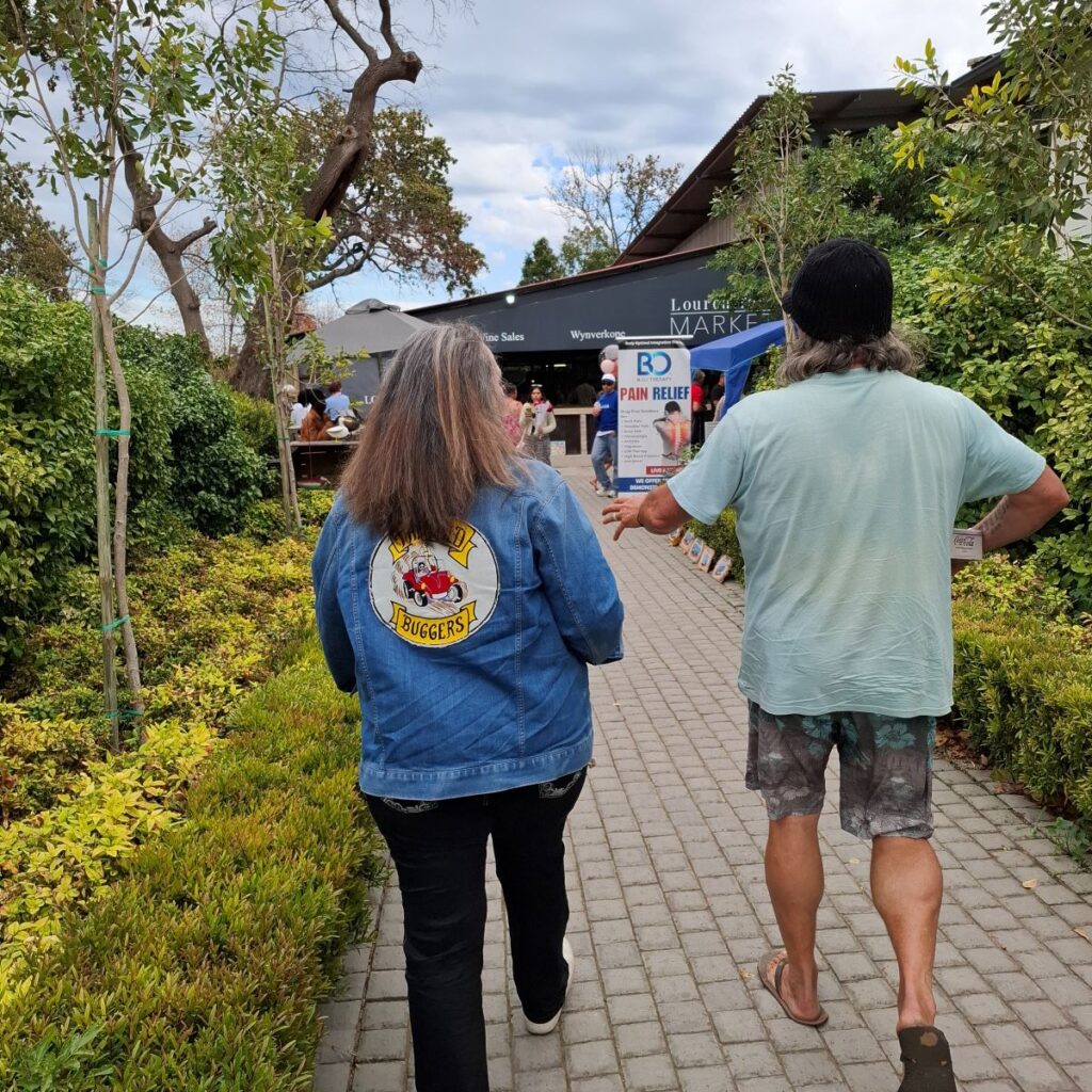 Mickey and Tony from the Wild Buggers Beach Buggy Club are seen walking towards the entrance of the Lourensford Estate restaurant area, with a beautiful garden or pathway visible around them, at the end of Wiehann's Run.