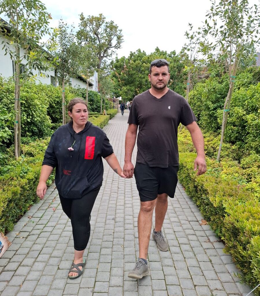 Lerene, with an unidentified fellow Wild Buggers Beach Buggy Club member, are walking towards the Lourensford Estate restaurant area, with the scenic setting and possibly other club members visible in the background, marking their arrival for lunch after Wiehann's Run in Cape Town.