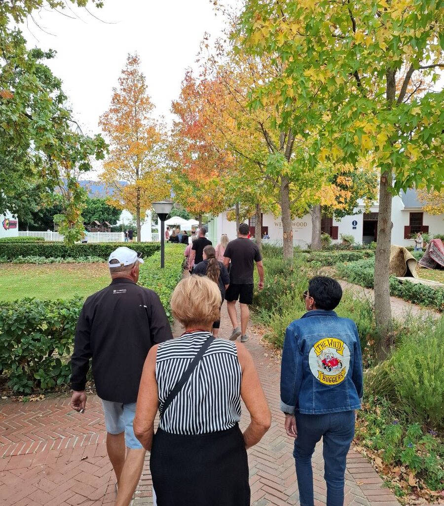 A larger group of Wild Buggers Beach Buggy Club members are walking towards the restaurant and garden area of Lourensford Estate in Cape Town, Western Cape, South Africa, indicating the final arrival of the group for their lunch stop at the conclusion of Wiehann's Run.