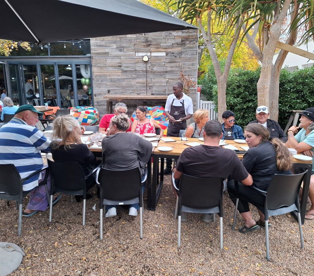 Wild Buggers Beach Buggy Club members are seated comfortably at a long table in the beautiful outdoor dining area of Lourensford Estate, relaxed and engaged in conversation, with menus visible, preparing to order food after their run.