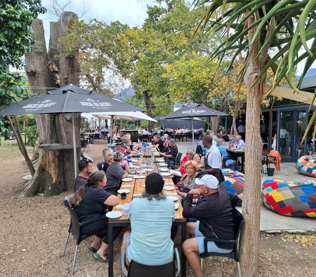 Wild Buggers Beach Buggy Club members are seated at a long table in the Lourensford Estate dining area, engaged in conversation, with some looking towards the kitchen or staff, indicating they are waiting for their food orders after Wiehann's Run.