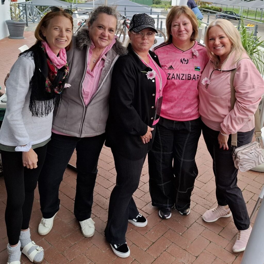 A group of women from the Wild Buggers Beach Buggy Club, dressed in black and pink clothing as a tribute, smiling and posing together at the Potbelly Pantry pit stop during Wiehann's Run.