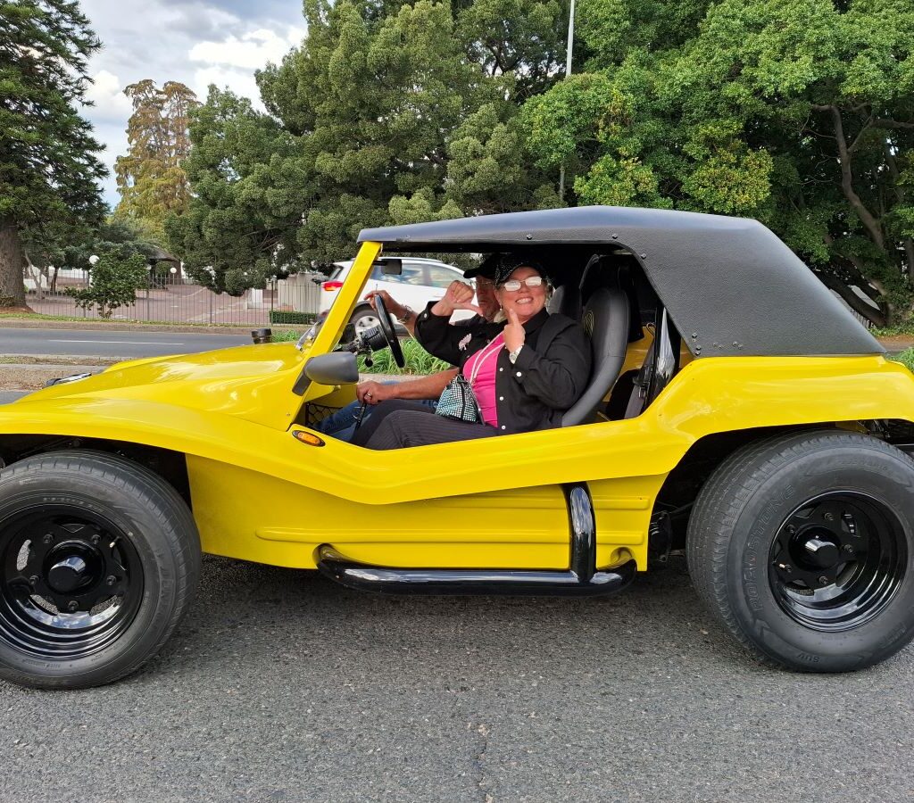 Anthony and Michelle Jenkins are seated in their yellow 1600 VW air-cooled Salamander beach buggy, "Bumble Bee," at Lourensford Estate, looking towards the camera with smiles, signifying the end of the Wild Buggers' Wiehann's Run.