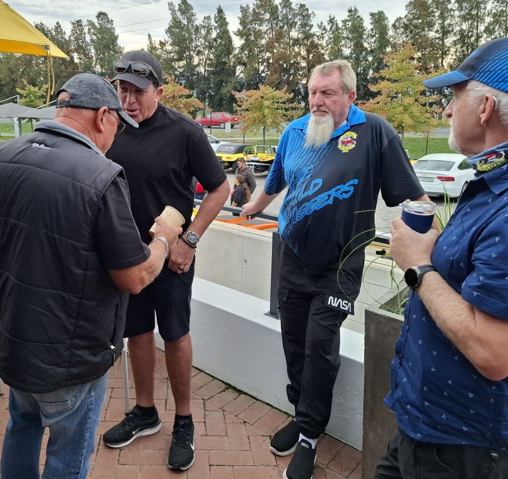 A group of Wild Buggers Beach Buggy Club men, all wearing something black as a tribute, standing and chatting on the balcony of Potbelly Pantry during the first pit stop of Wiehann's Run, with casual camaraderie evident.