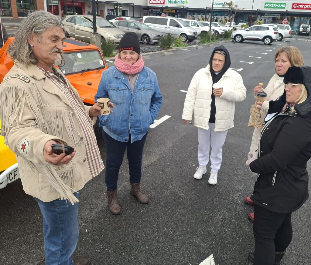 A group of Wild Buggers in a wet parking lot at Bothasig, with their Beach Buggies and VW Air-cooled vehicles visible in the background, listening intently to Tony.