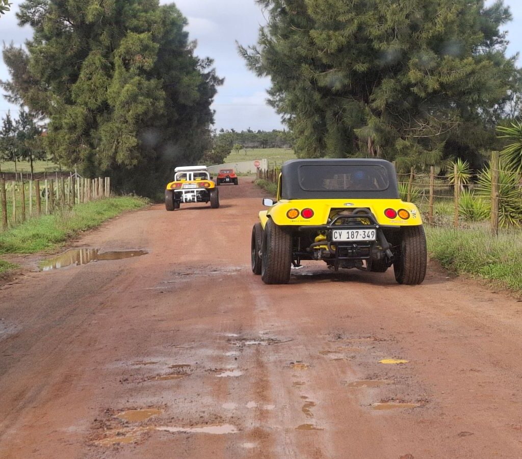 Another perspective of Wild Buggers in their Beach Buggies and VW air-cooled vehicles driving along a wet gravel road towards the "African Savanna."