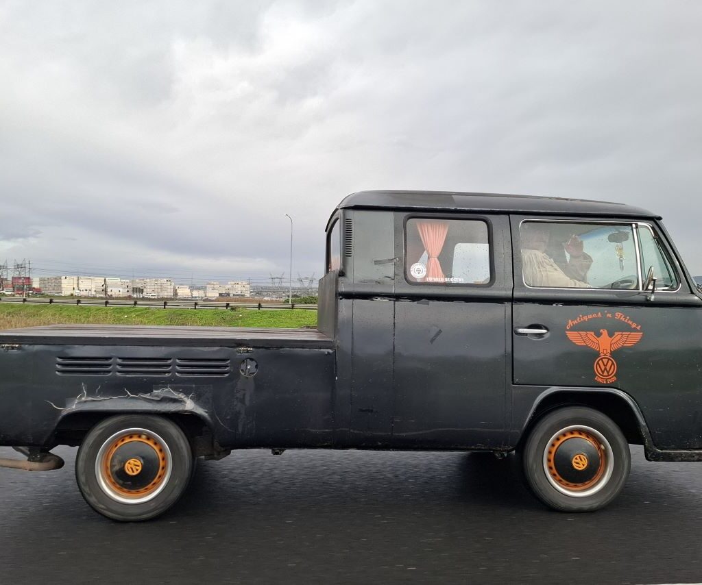 Anton and Mickey in their Baja Bug driving alongside Tony in his VW air-cooled Kombi on a road during The Wild Buggers club run, with Anton taking a photo of the Kombi.