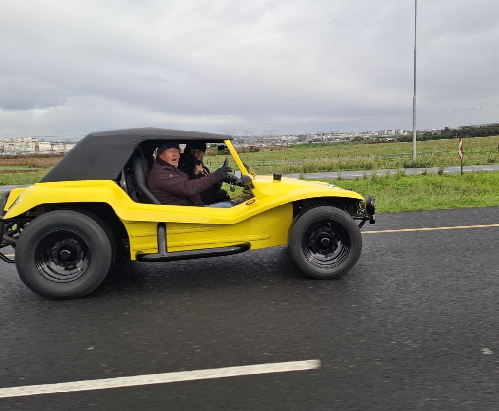 Anton and Mickey in their Baja Bug (unseen) on a road, taking a photo of Anthony and Michelle Jenkins in their bright yellow VW air-cooled Salamander Beach Buggy from the driver's side perspective.