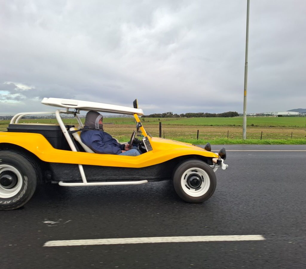 Anton and Mickey in their Baja Bug (unseen) on a road, taking a photo of Chris and Sandra Zara in their bright yellow VW air-cooled Salamander Beach Buggy from a side perspective.