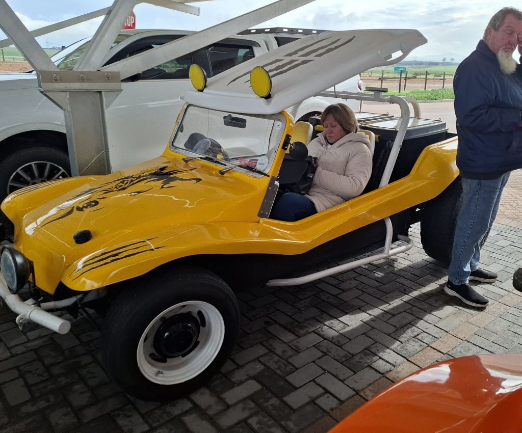 Chris standing next to his Beach Buggy in a parking area while Sandra sits inside the vehicle, during The Wild Buggers Tony & Sue's Run.