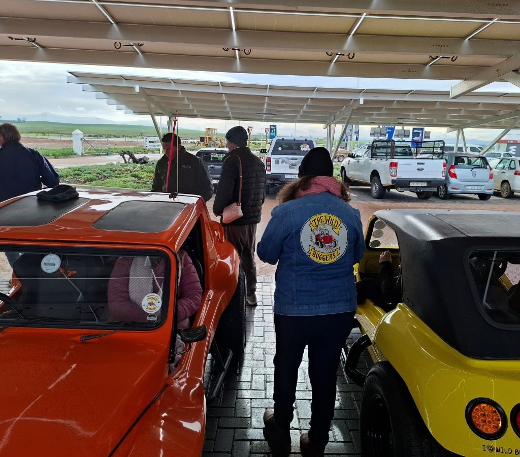 Mickey and Sam from The Wild Buggers Beach Buggy Club having a relaxed conversation in a parking area, with their Baja Bugs visible nearby.