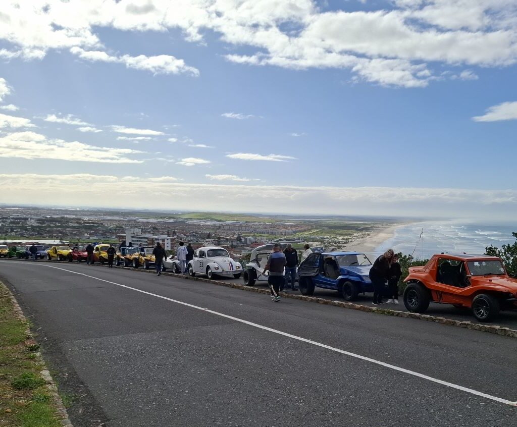 Long line of colorful VW classics and beach buggies parked at a pitstop with ocean in background during Wild Buggers 'Revving Up for the Lens' cruise
