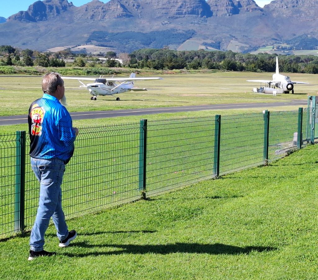 A photo of Wild Buggers Beach Buggy Club member Chris Zara standing behind a fence next to an airstrip at the Stellenbosch Flying Club, looking at a vintage fighter jet and several small private planes on a sunny day.