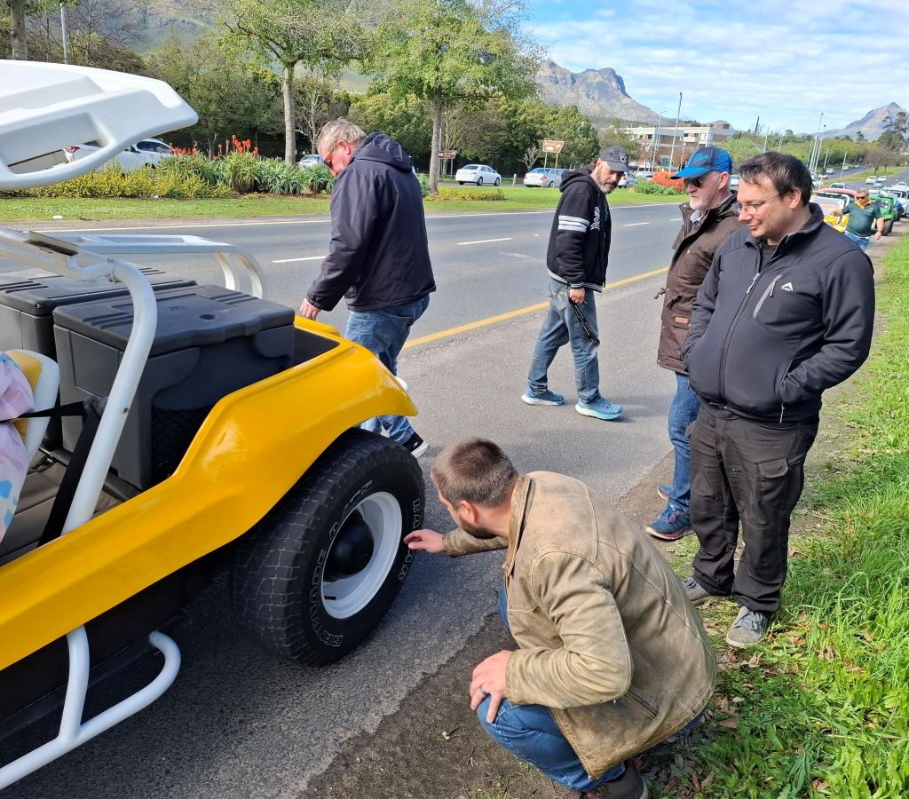 A photo of several Wild Buggers' club members gathered around Chris Zara's yellow Salamander beach buggy, "Zara's Bug," during an emergency pitstop, looking at the wheel to diagnose a problem on the side of the road. Zara's Bug has a 1600 VW air-cooled Beetle motor.