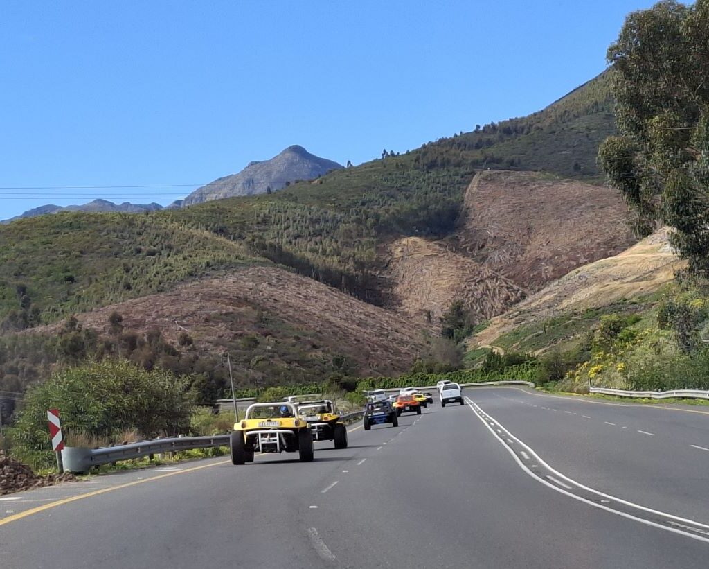 A photo of a convoy of Wild Buggers' beach buggies and VW Beetles driving on a winding road towards Hellshoogte Pass, with a backdrop of mountains.