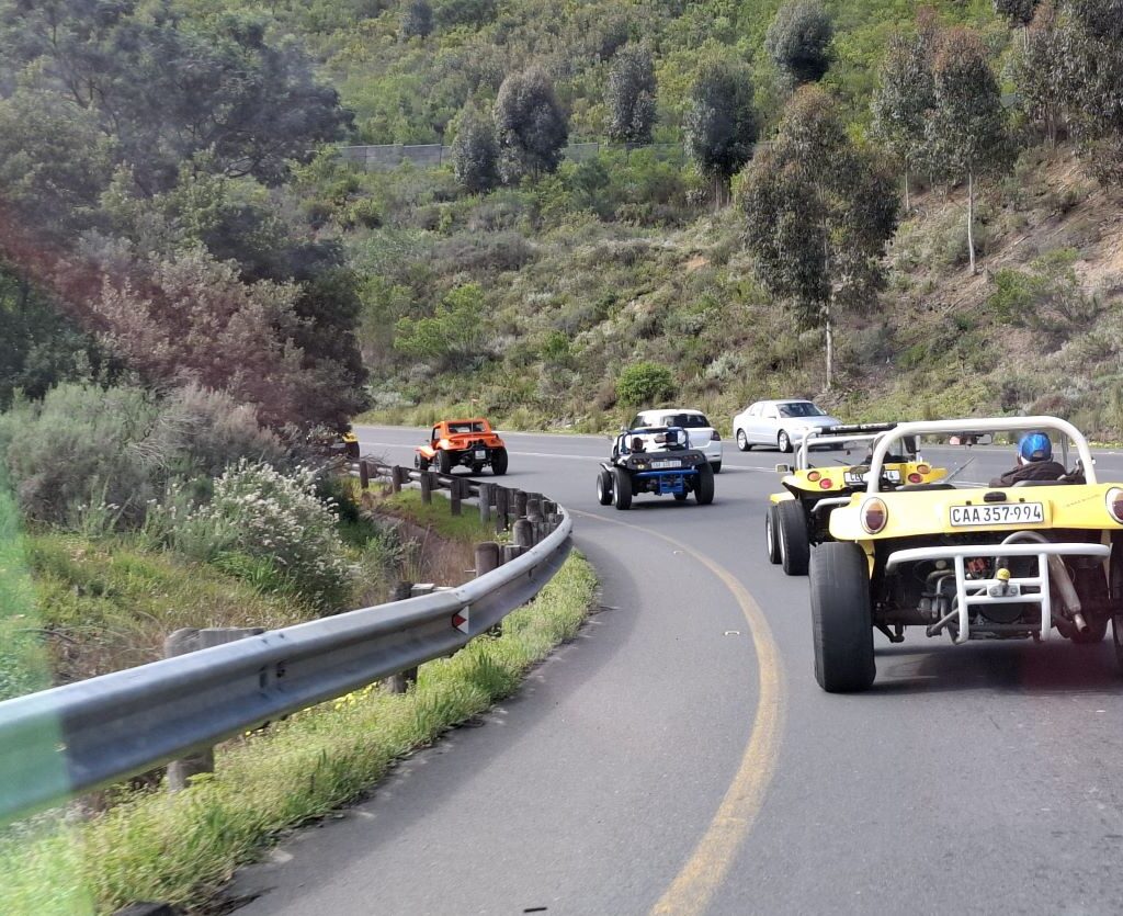 A photo of a convoy of Wild Buggers' beach buggies and VW Beetles continuing their drive up a winding mountain road, surrounded by lush green foliage and trees.