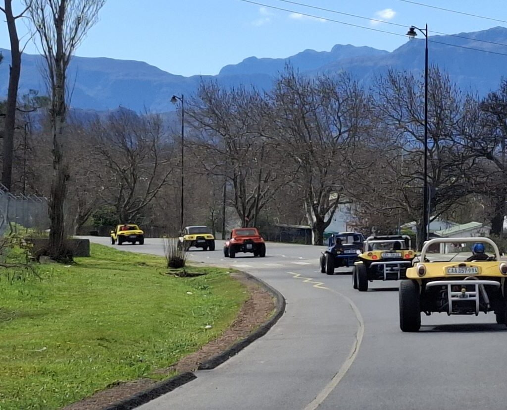 A photo of a convoy of Wild Buggers' beach buggies and VW Beetles entering a small town, driving on a quiet road with a few buildings in the background.