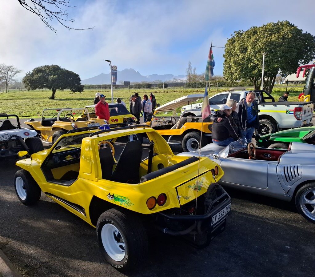 A close-up shot of several Wild Buggers' beach buggies, VW Beetles, and other VW classics gathered at the meeting point on a sunny Sunday, with club members chatting before the start of Nick & Ellie's Run.