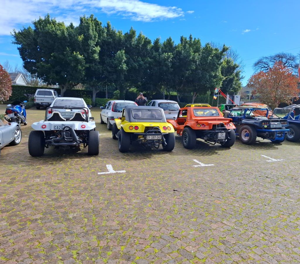 A photo of a lineup of Wild Buggers' vehicles, including beach buggies, VW Beetles, and other VW classics, parked neatly in the parking lot of the Franschhoek Beer Company.