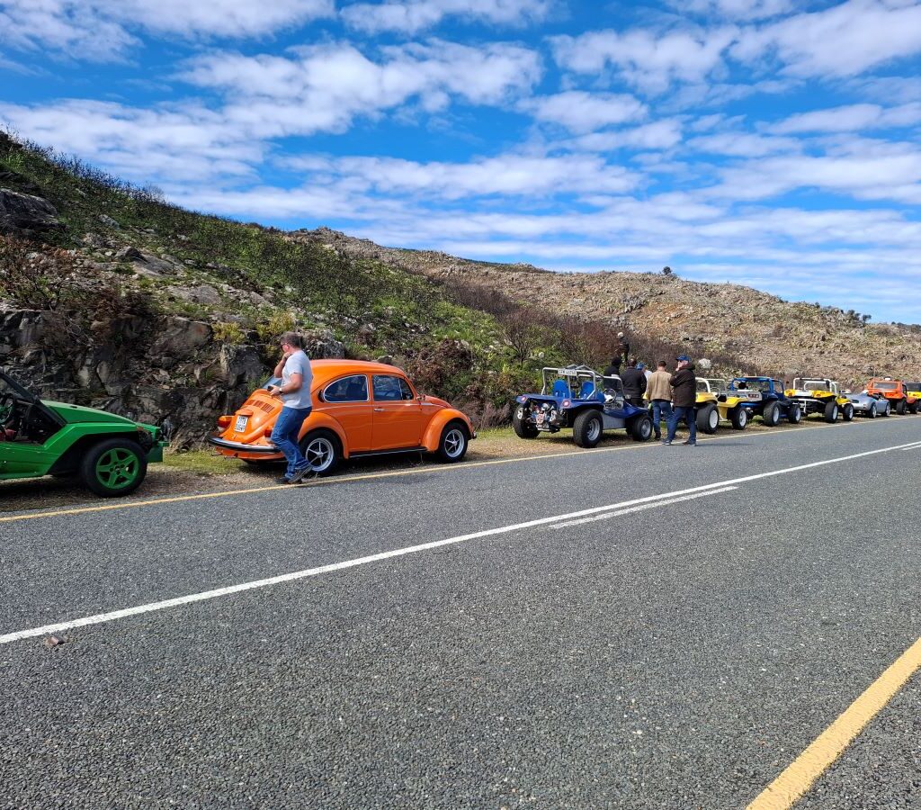 A photo of several Wild Buggers' beach buggies and VW Beetles parked on the side of a road during a pitstop, with a beautiful scenic view of the surrounding Cape landscape. Club members are standing around their vehicles, chatting.