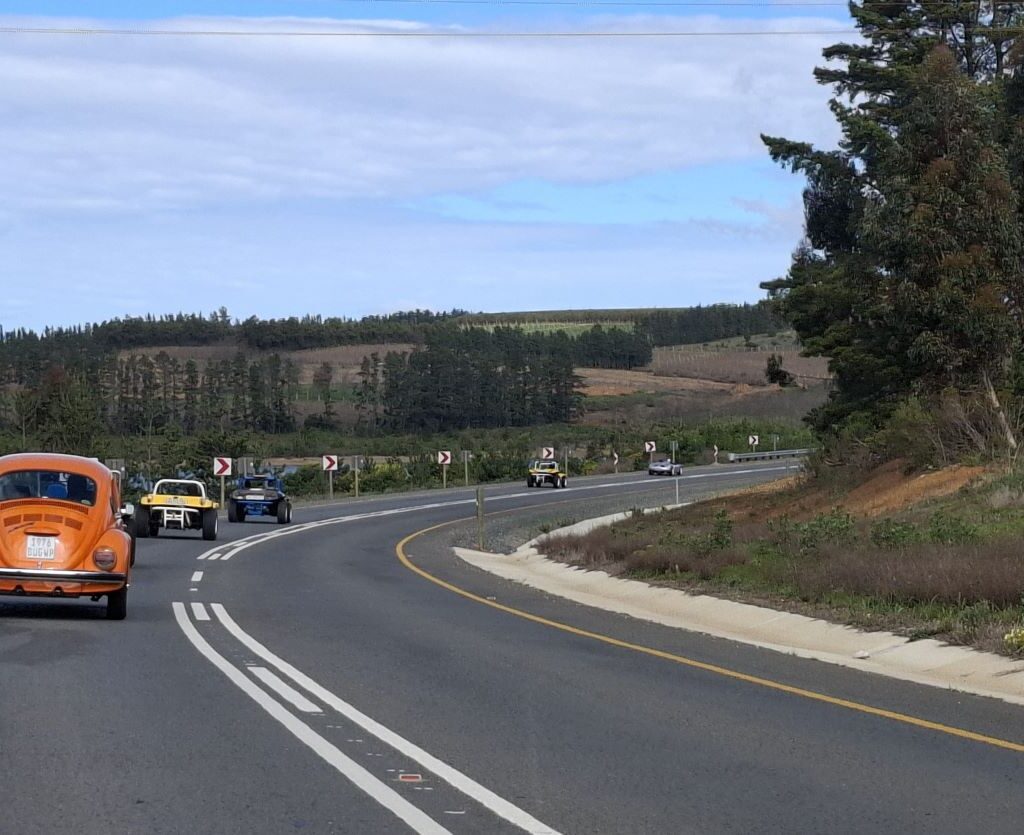 A photo of a convoy of Wild Buggers' vehicles, including beach buggies and VW Beetles, driving on a winding road with mountains and green scenery in the background.