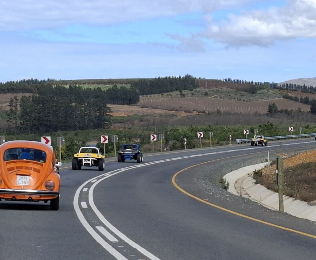 A photo of a convoy of Wild Buggers' vehicles, including beach buggies and VW Beetles, driving on a highway with open, green fields in the background.