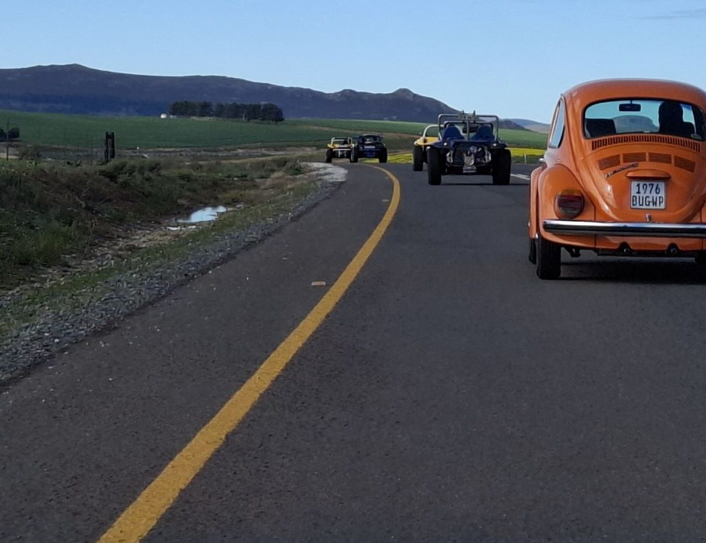 A photo of a convoy of Wild Buggers' vehicles, including beach buggies and VW Beetles, driving on a road, continuing their journey with mountains and a green landscape in the background.