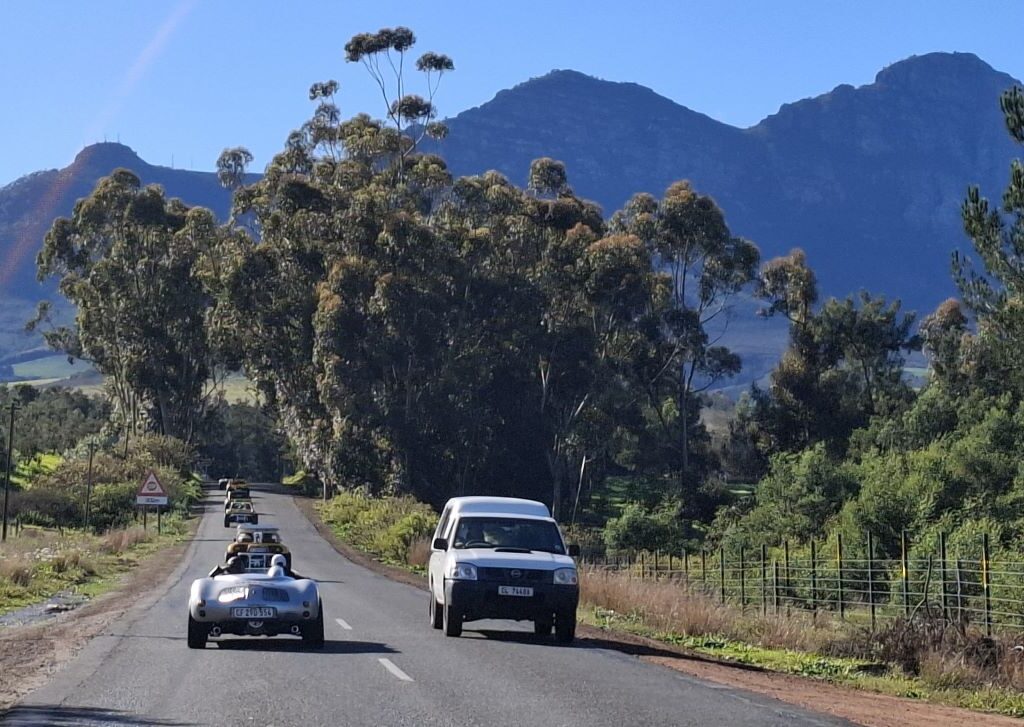 A photo of a convoy of Wild Buggers' beach buggies and VW Beetles driving on a road towards Franschhoek, with beautiful mountains visible in the foreground.