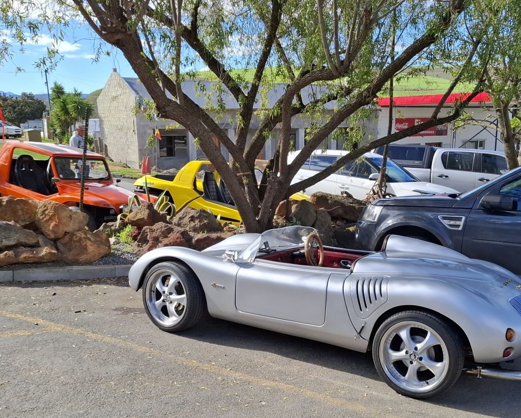 A photo of several Wild Buggers' vehicles, including beach buggies and VW Beetles, parked in the parking lot of the Botrivier Hotel.