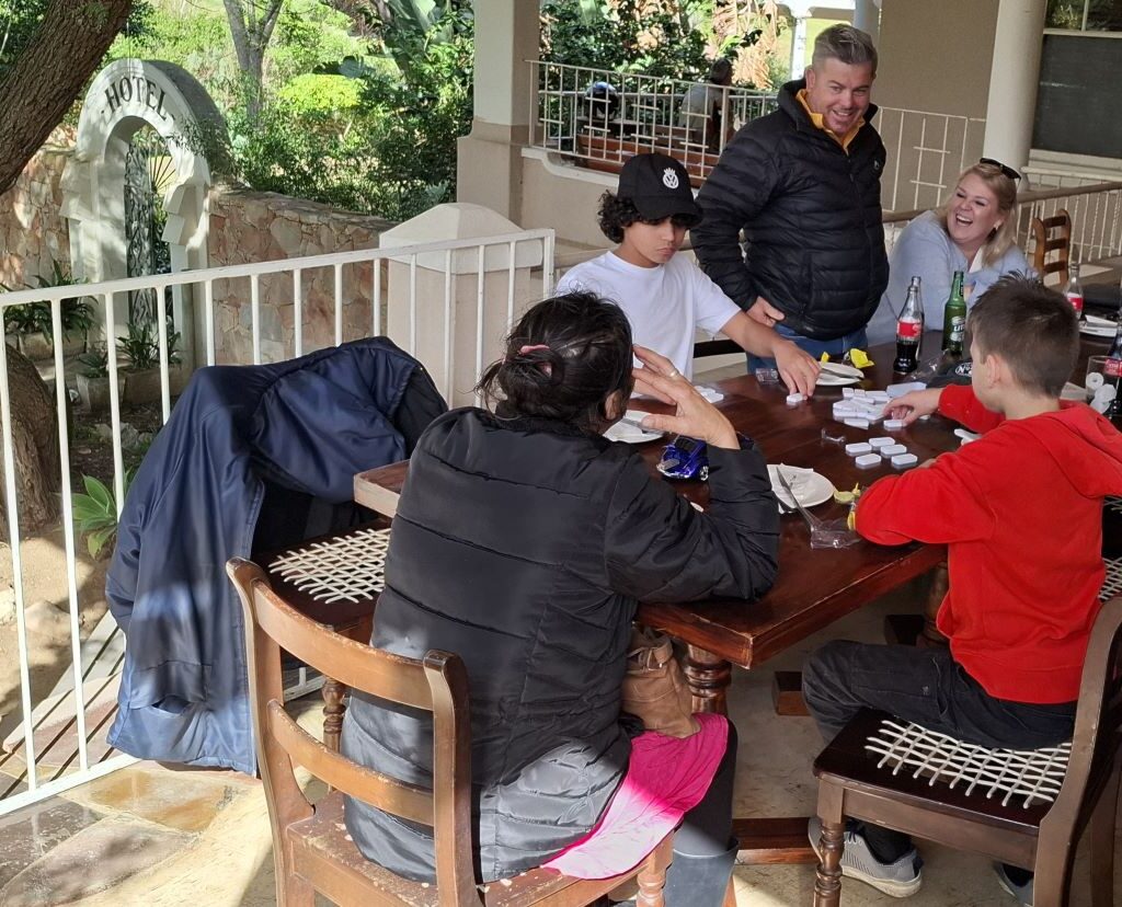A photo of several Wild Buggers' club members sitting at a table at the Botrivier Hotel, laughing and playing a game of dominoes while waiting for their lunch.