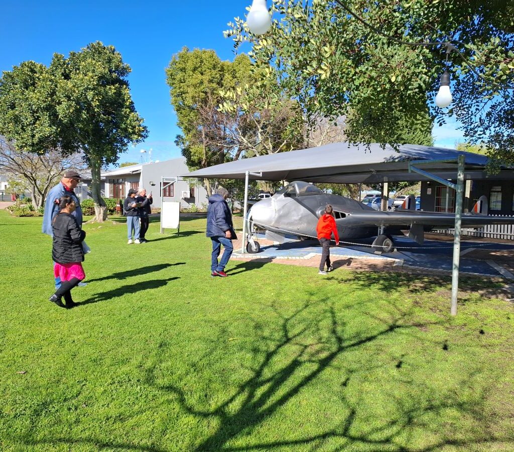 A photo of a vintage South African fighter plane on display at Stellenbosch Flying Club, with several Wild Buggers Beach Buggy Club members walking around and enjoying the scenery in the background.